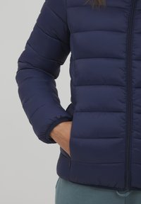 Námornícky modrá zateplená bunda so zipsom vpredu, horizontálnym prešívaním a elastickými manžetami. Obsahuje bočné vrecká s elegantným dizajnom.