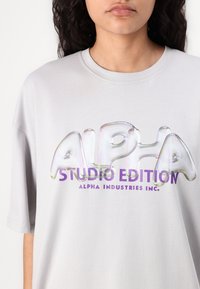 T-shirt en coton gris surdimensionné arborant un grand graphique en relief brillant "ALPHA" aux couleurs iridescentes avec "STUDIO EDITION" en violet en dessous.