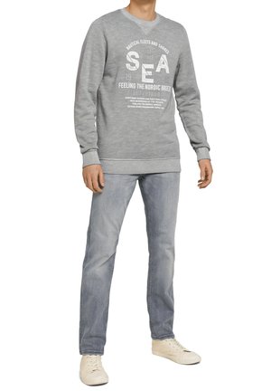 Grauer Pullover mit runder Halslinie, gerippten Bündchen und Saum. Mit weißem Drucktext und Grafiken. Kombiniert mit hellgrauen Jeans und beigen Schuhen.