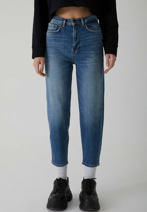 Light blue denim jeans with a high waist, tapered leg, and slight fading. Worn with black sneakers and white socks, revealing a cropped top.