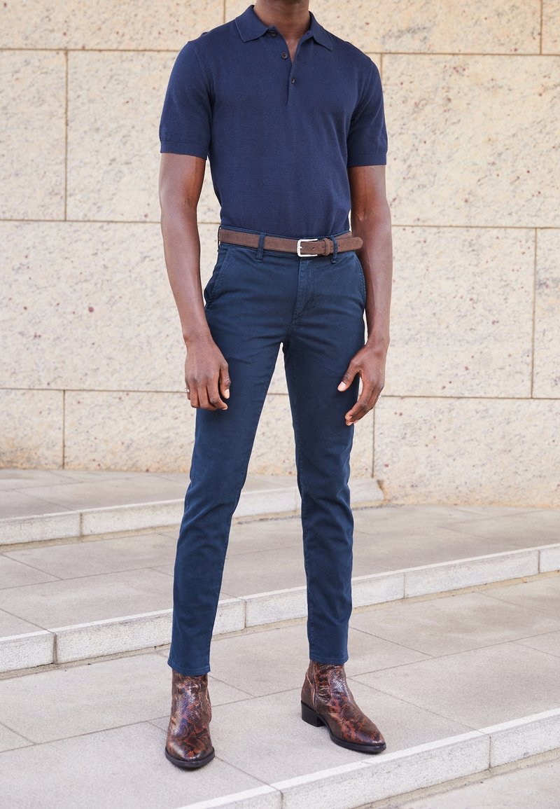 Homme debout sur des marches, portant un polo bleu marine, un pantalon bleu foncé, une ceinture marron et des bottines marron motif peau de serpent.