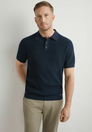 Mężczyzna z krótkimi włosami i brodą, ubrany w granatową prążkowaną koszulkę polo i beżowe spodnie, stoi na tle jasnej, gładkiej ściany.