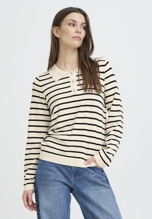 Fiatal nő krémszínű, hosszú ujjú, fekete csíkos galléros pólót és farmernadrágot visel, egy kézzel a zsebében áll.