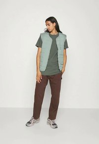 Mlad človek z dolgimi temnimi lasmi, oblečen v svetlo zeleno podloženo jopico preko temno zelene majice, rjavih hlač in belih superg, stoji sproščeno.