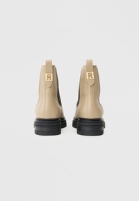 Bottines en cuir beige avec des panneaux élastiques noirs sur les côtés, semelles épaisses noires et accents en métal doré sur les languettes à l'arrière.