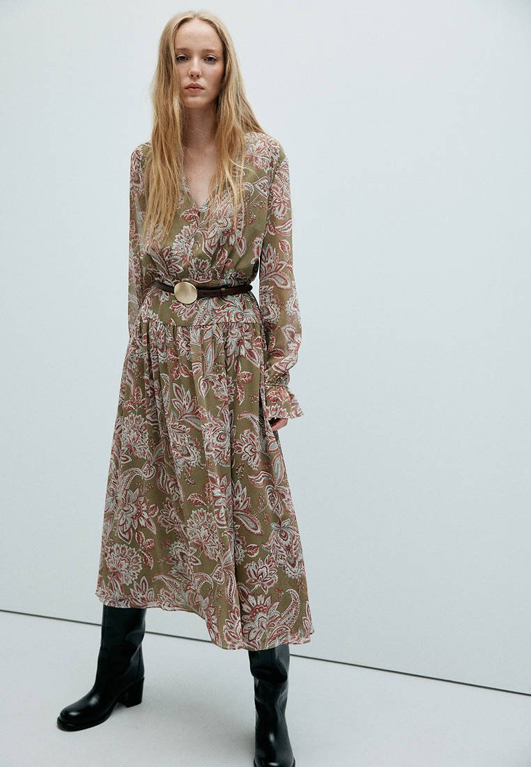 Femme aux longs cheveux blonds portant une robe verte à fleurs à manches longues avec une ceinture noire et des bottes noires montantes, debout devant un fond uni.