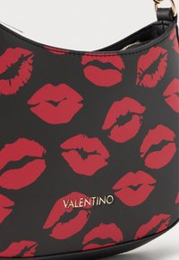 Crna torba sa crvenim otiscima usana, s teksturiranom površinom i zlatnim logotipom "Valentino". Ima zakrivljen oblik i malu naramenicu.