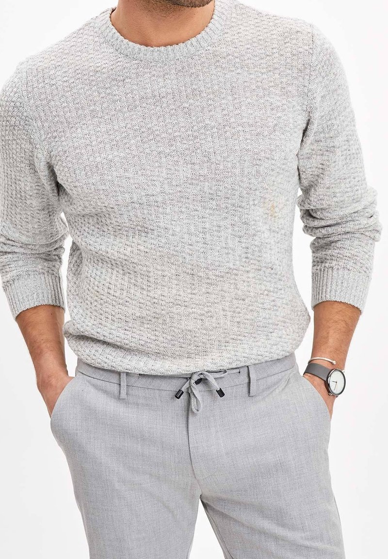 Homme portant un pull en maille texturée gris clair et un pantalon gris clair assorti, les mains dans les poches, montre visible au poignet gauche.