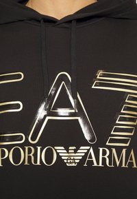 Svart hooded sweatshirt med en stor guld grafisk logotyp med metallisk finish, som visar texten "EA7 EMPORIO ARMANI." Detaljerna inkluderar varierande bokstavshöjder.