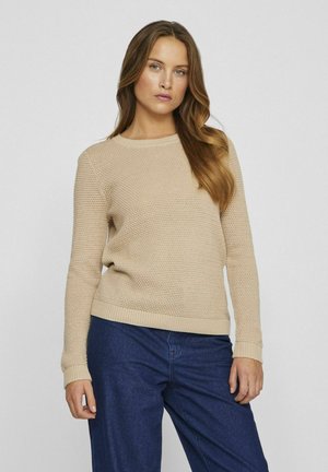 Pull en tricot beige avec un motif texturé, col rond et bords côtes aux poignets et à l'ourlet, associé à un pantalon en denim bleu foncé.