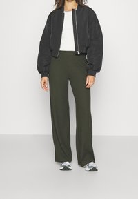 Svart bomberjacka med puffärmar, vit åtsittande skjorta, olivgrön ribbade vida byxor, och silverfärgade träningsskor. Slät textur och enkel design.