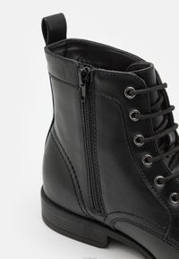 Botín negro de cuero hasta el tobillo con cremallera lateral, cordones en la parte delantera, puntera redonda y un acabado texturizado con detalles de hardware plateado.