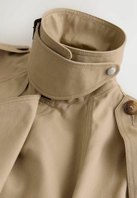 Khaki trenchcoat med høj krave, der har lukning med knapper og glat, tekstureret stof. Detaljer inkluderer kontrasterende knapper og sømme.