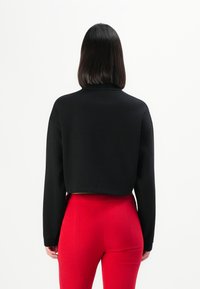 Person med skulderlangt mørkt hår iført en sort langermet crop-top og høy midje røde bukser, vist fra bak.