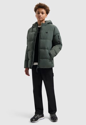 Gevoerde groene jas met een ritssluiting aan de voorkant, capuchon en zwarte accenten; gedragen over een zwarte trui, gecombineerd met zwarte broek en sneakers.