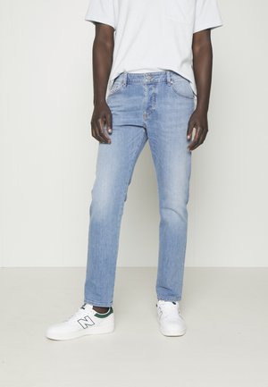 Osoba nosząca jasnoniebieskie dżinsy, prostą białą koszulkę i białe trampki New Balance stoi przy białej ścianie.