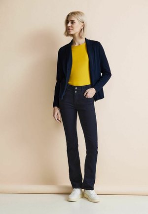 Blazer marine sur un haut jaune, associé à un jean foncé et des baskets blanches. Le blazer a une coupe ajustée et un subtil détail de bouton.