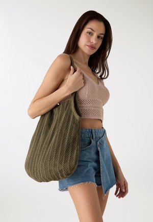 Zielona siatkowa torba z fakturowaną powierzchnią, wyposażona w szerokie otwarcie i solidne uchwyty. Modelka nosi dziergany top i jeansowe szorty.