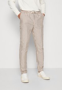 Pantaloni a righe beige con strisce verticali blu, vita con cordino e gamba affusolata, abbinati a sneaker bianche. Materiale in cotone morbido.