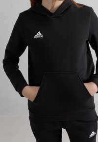 Sudadera negra con capucha hecha de tela suave, con un bolsillo tipo canguro y el logo blanco de Adidas en el lado izquierdo del pecho.