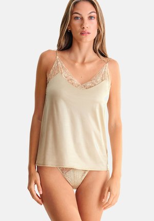 Femme portant un caraco en satin beige avec des bordures en dentelle et une lingerie assortie en dentelle, debout devant un fond clair uni.