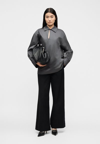 Γυναίκα με γκρι σκούρο φαρδύ μπλουζάκι, μαύρο παντελόνι με φαρδιά μπατζάκια, μαύρες μυτερές γόβες, κρατώντας μεγάλη μαζεμένη μαύρη δερμάτινη τσάντα φάκελο.