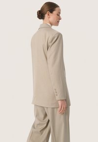 Beige szabott blézer egyetlen hátventilátorral, két gombbal az ujjakon, struktúrált vállakkal, illesztve az azonos színű, bővített nadrággal.