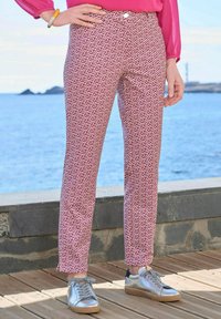 Geprinte broek in een rood en wit geometrisch ontwerp, slim fit, gecombineerd met zilveren sneakers. Achtergrond toont een uitzicht op het water.