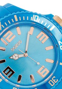 Montre en silicone bleue avec un boîtier rond. Cadran bleu clair, index horaires en rose doré et fenêtre de date à 6 heures. Lunette texturée et accents métalliques.