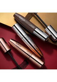 Trois tubes de mascara KIKO Milano en marron, argent et or rose, avec des finitions brillantes et des applicateurs noirs. Présentés sur un fond rouge et or.