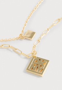 Halskæde i guldton med en firkantet vedhæng med udskårne "DKNY"-bogstaver, der kombinerer led- og kædedesign for tekstur og visuel interesse.
