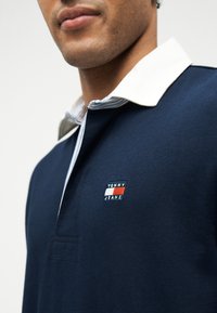 Κοντινό πλάνο ενός ατόμου που φοράει μπλε πουκάμισο πόλο με λευκό γιακά και λογότυπο Tommy Jeans στο στήθος.