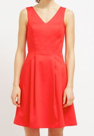 Ärmelloses rotes Kleid mit V-Ausschnitt und figurbetontem Oberteil, das sich zu einem ausgestellten A-Linien-Rock erweitert, getragen von einer stehenden Person vor einfachem Hintergrund.