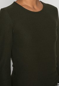 Nahaufnahme einer Person, die ein dunkelgrünes, geripptes Langarmshirt mit rundem Ausschnitt vor einem schlichten Hintergrund trägt.