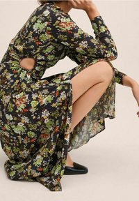 Květinové černé šaty s dlouhými rukávy, se zvýrazněním v oblasti pasu. Textilie zobrazuje barevné květiny v zelené, oranžové a bílé.