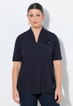 Kvinde med mørkt hår i en glat knold bærer en marineblå kortærmet top med en blomsterbrosje og perleophængsøreringe mod en hvid baggrund.