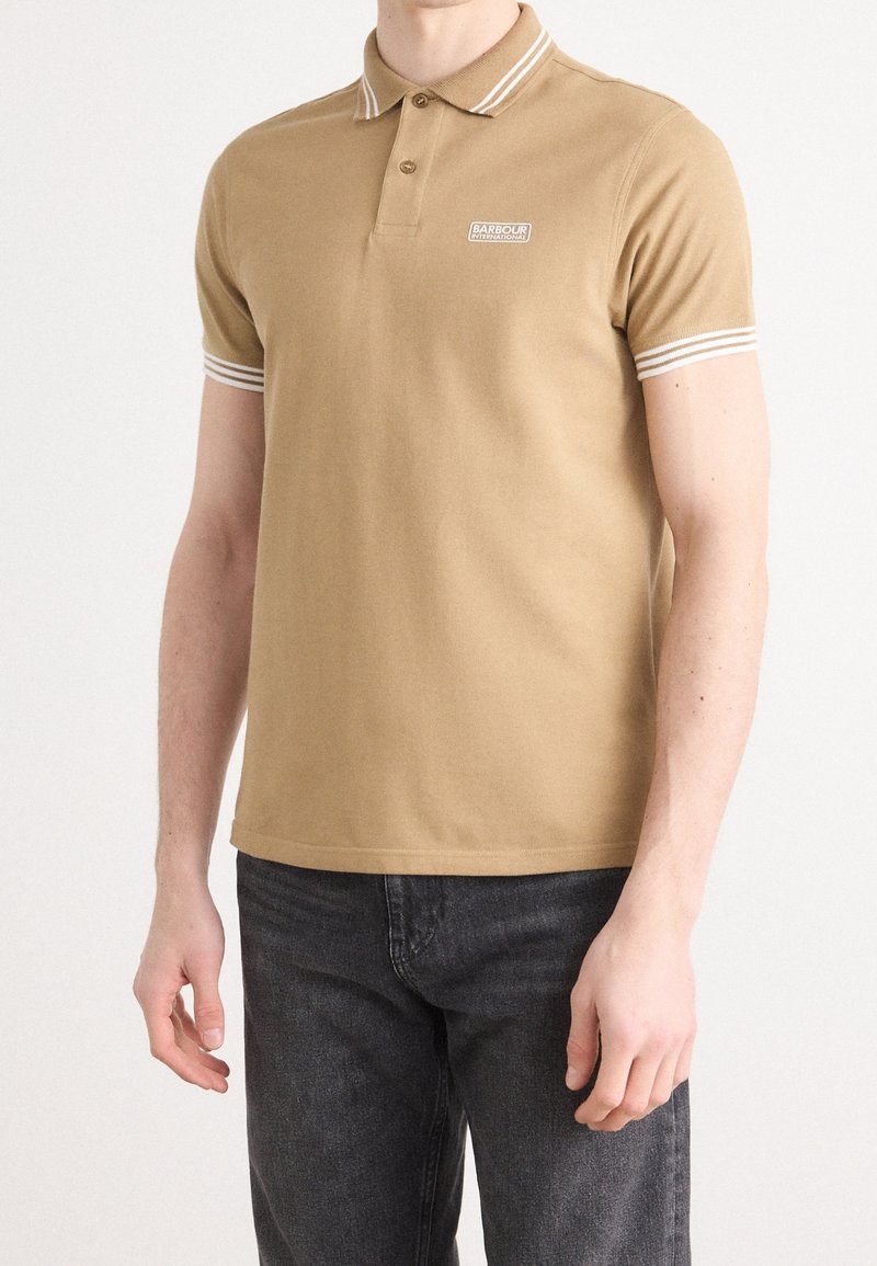 Homme portant un polo à manches courtes couleur beige avec des rayures blanches sur le col et les manches, associé à un jean noir, debout devant un fond uni.