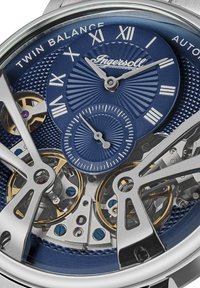 Automatyczny zegarek z niebieską teksturowaną tarczą, srebrzystą kopertą, widocznymi detalami mechanizmu oraz złotymi akcentami, z oznaczeniami godzin w postaci cyfr rzymskich.