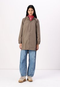 Lichtbruine, waterdichte jas met een capuchon en elastische manchetten, gecombineerd met losse blauwe jeans en beige plateau schoenen. Rode bandana als accent.