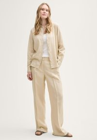 Camicia di lino beige e pantaloni a gamba larga, entrambi di vestibilità comoda, abbinati a una canotta bianca e sandali bassi con una fascia testurizzata.