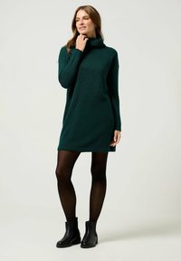 Abito verde con collo alto, maniche lunghe, tessuto strutturato e spacchi laterali, abbinato a stivali neri alla caviglia e collant neri trasparenti.