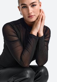 Top lung cu mâneci lungi din material transparent, negru, cu o textură subtilă, asortat cu pantaloni din piele ecologică negri. Mâinile modelului sunt unite aproape de față, pe un fundal neutru.