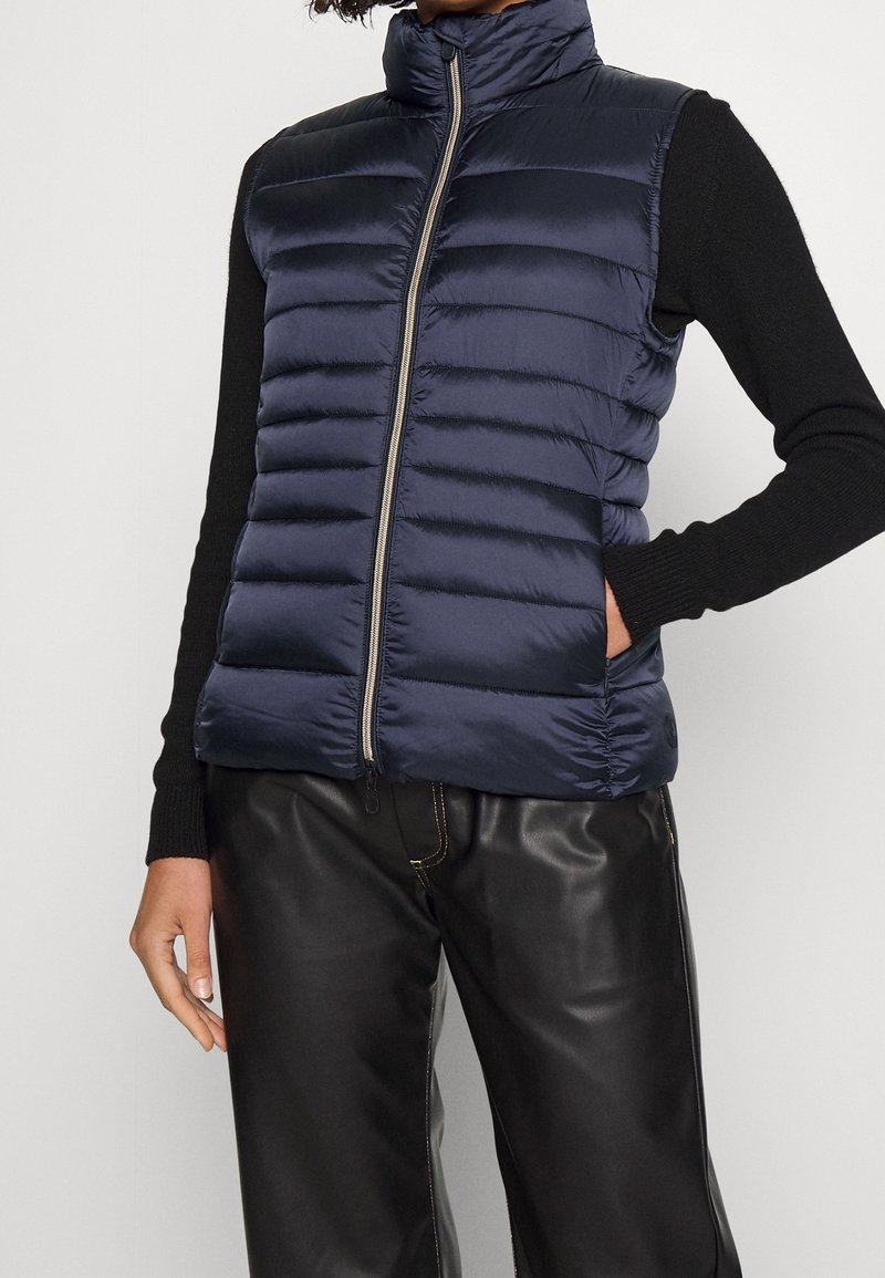 Marineblauer Steppwesten mit horizontalem gestepptem Design, hohem Kragen, Frontreißverschluss und Seitentaschen, getragen über einem schwarzen Langarmoberteil.