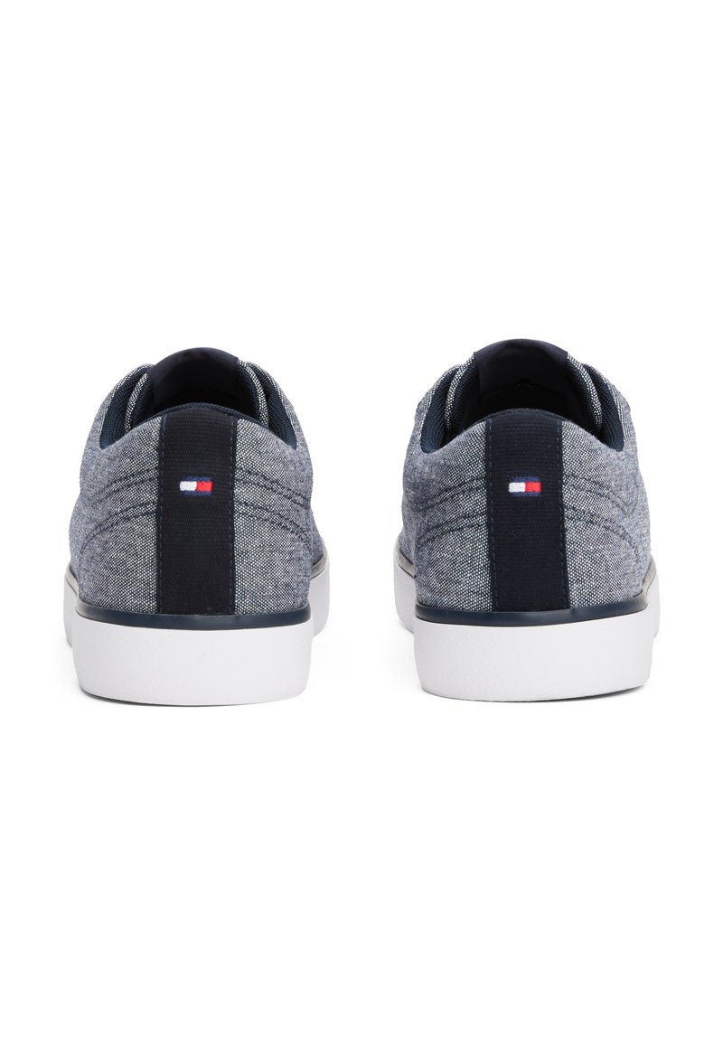 Baskets en tissu gris avec une finition texturée, semelles en caoutchouc blanches et accents bleu marine. Présente un petit logo rouge, blanc et bleu sur le talon.