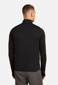 Uomo di spalle che indossa un gilet senza maniche nero sopra una maglietta a maniche lunghe nera e pantaloni grigi su uno sfondo bianco uniforme.