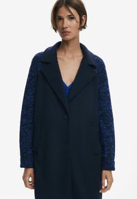 Cappotto in lana blu navy con maniche in maglia blu contrastante, dotato di colletto a punte, chiusura con bottoni e tasche laterali. Tessuto liscio e forma strutturata.