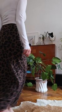 En person i en hvit langermet topp og skjørt med leopardmønster står nær et tre-skap med planter, lys og dekorative gjenstander.