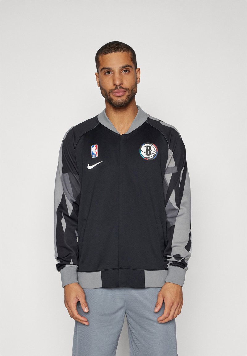Black and grey basketball jacket with a zip front, a ribbed collar, and team logo on chest. Features geometric patterns on sleeves.