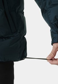 Chaqueta acolchada en azul marino oscuro, con una textura suave y brillante. Una mano tira de un cordón ajustable en el dobladillo para un ajuste más ceñido.