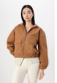 Veste zippée marron en matériau doux avec deux poches avant, ourlet élastique et col haut, assortie d'une chemise beige et d'un pantalon de couleur claire.